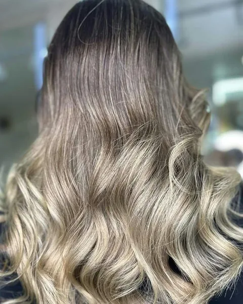 Long, glossy hair with dark roots melting into creamy blonde balayage, styled in soft waves